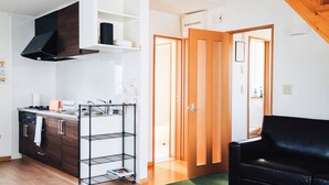 A modern living room with a black sofa, a kitchen area with a built-in stove, and a staircase with wooden beams. - Trailside Apartments & Chalet (Kutchan)