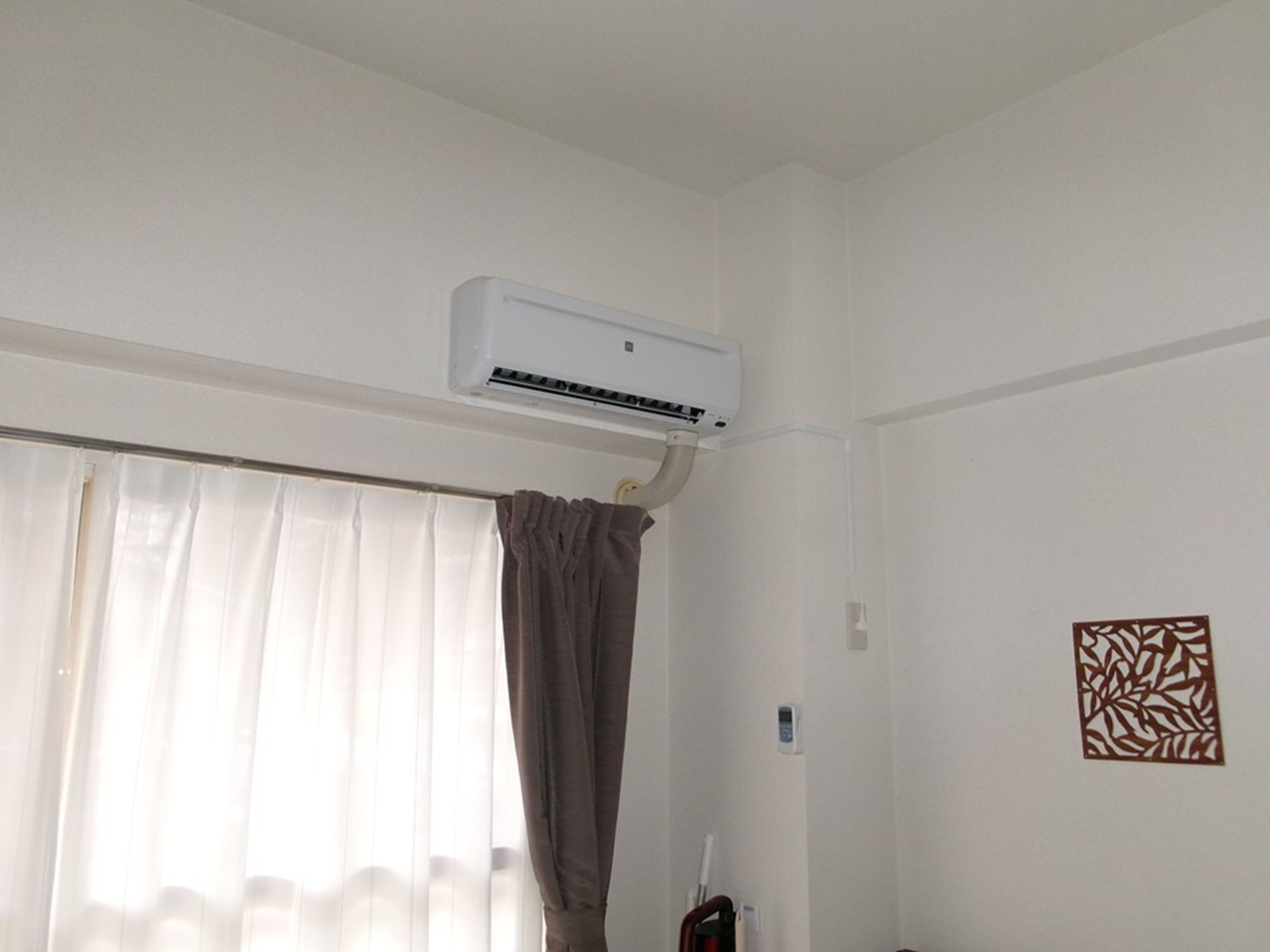 Une pièce dans laquelle un climatiseur blanc est fixé au mur, au-dessus d’une fenêtre ornée de rideaux blancs et légers.