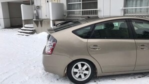 A beige sedan parked in front of a building with a white exterior, a set of steps leading to a door, and a window with a curtain.
