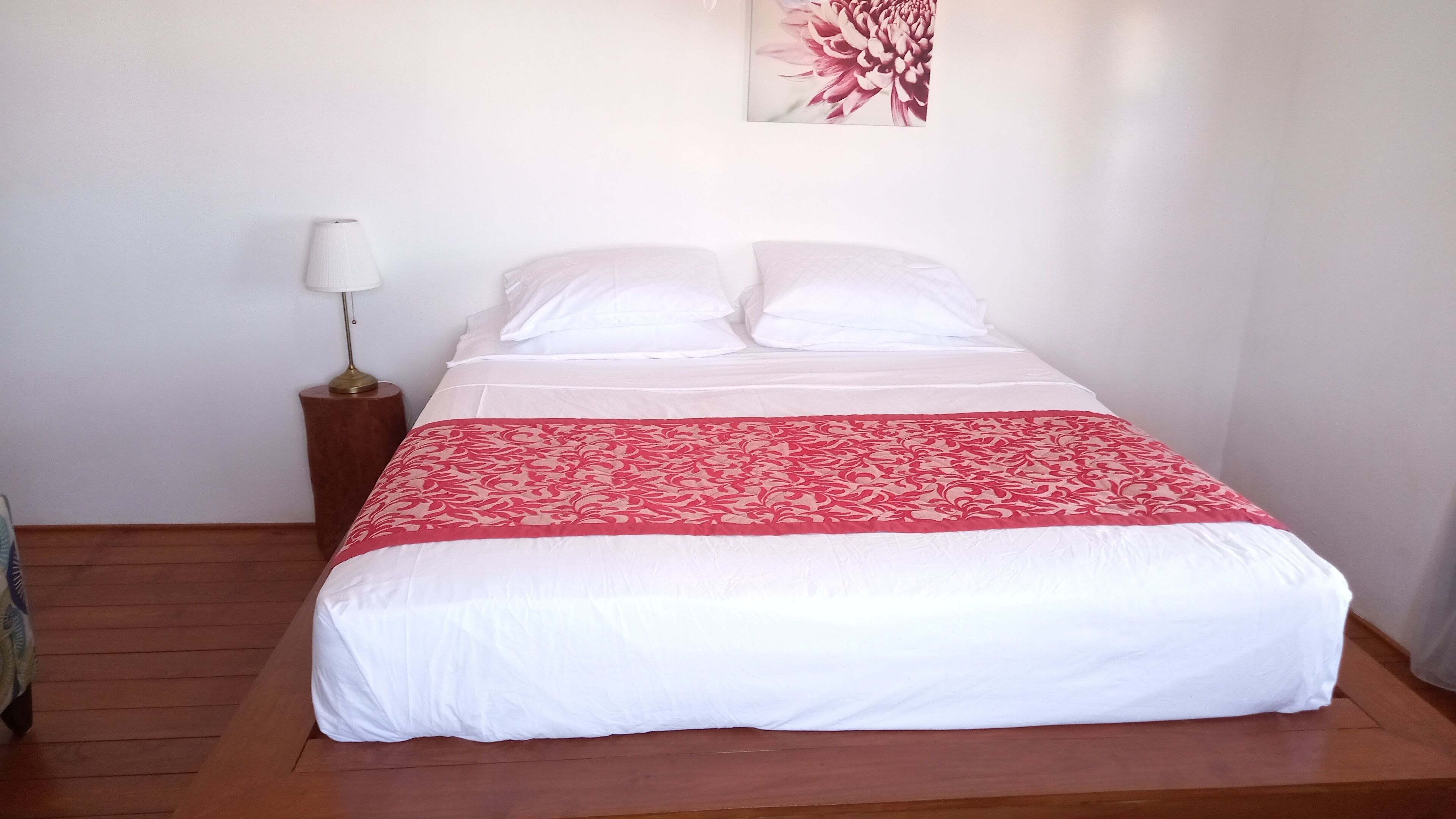 Een net opgemaakt bed met witte lakens en een rode geborduurde plaid, een bedlampje en een bloemig kunstwerk aan de muur.