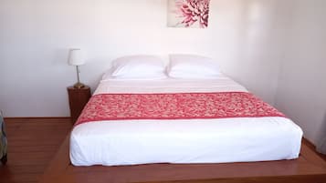Een net opgemaakt bed met witte lakens en een rode geborduurde plaid, een bedlampje en een bloemig kunstwerk aan de muur.