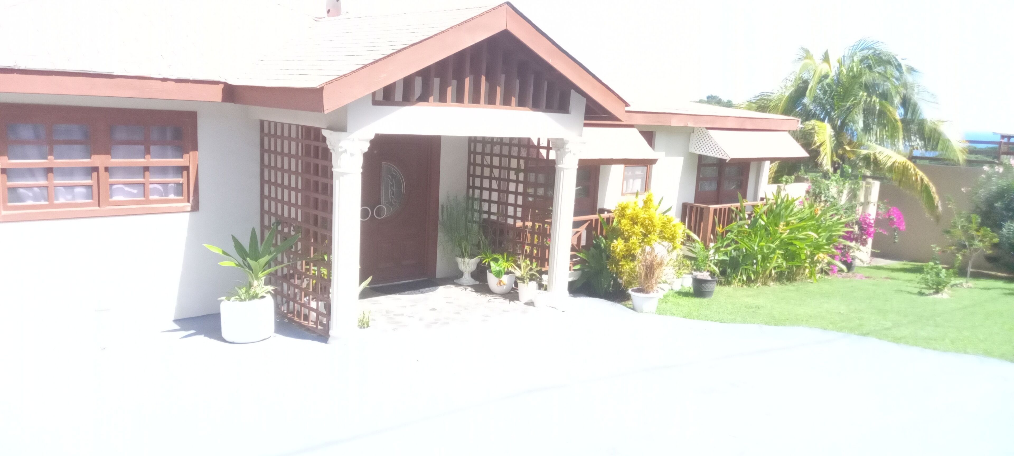 A house with a white exterior, a brown roof, and a wooden porch. There are potted plants and flowers in front of the house.