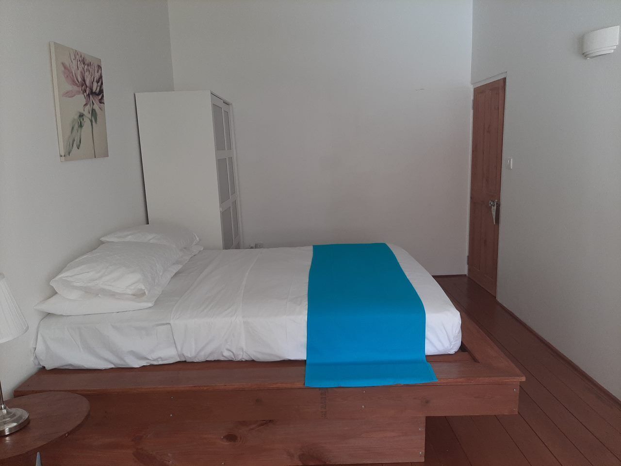 Una cama individual con ropa de cama blanca y un detalle azul, una mesita de noche de madera con una lámpara, y una puerta a la derecha.