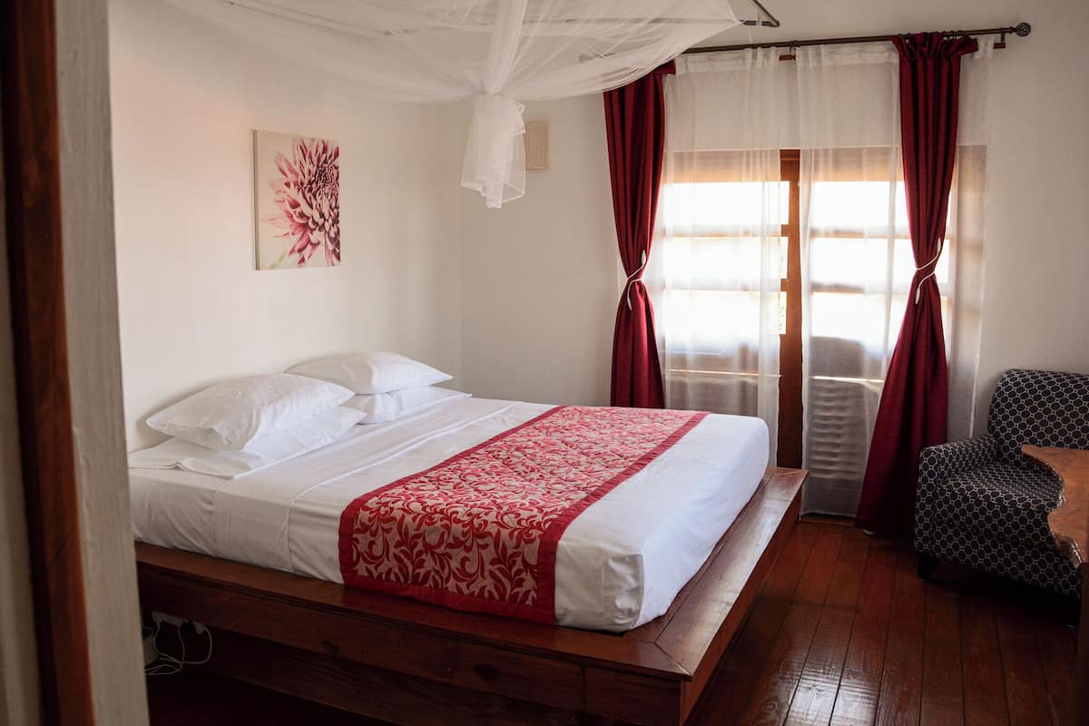 A neatly made bed with white linens and a red patterned bedspread, a wooden nightstand, a chair with a geometric pattern, and a window with red curtains.