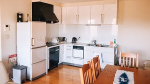 Uma cozinha compacta com armários brancos, geladeira, lava-louças e fogão. Há uma mesa de jantar com cadeiras e uma pequena mesa lateral.