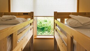 Duas beliches de madeira com roupa de cama branca, uma janela com vista para vegetação e um radiador na parede.