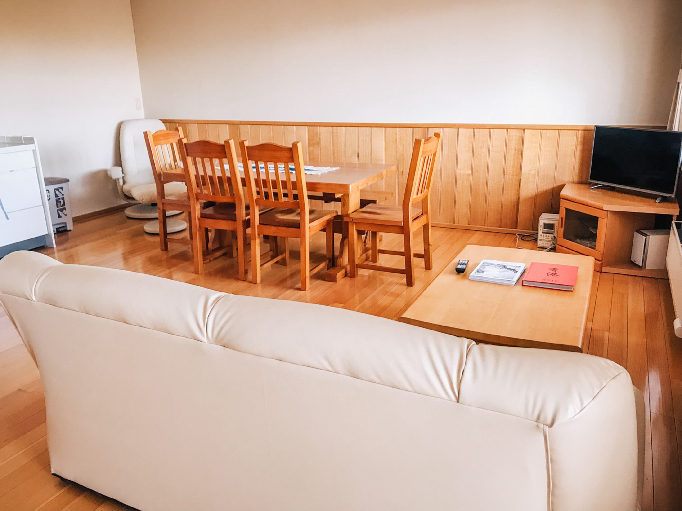 A cozy living room with a wooden dining table, four wooden chairs, a beige sofa, a small TV on a wooden stand, and a few books on the table.