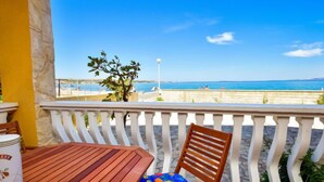 A wooden table and a chair with a colorful cushion on a balcony with a white railing, overlooking a beach and the sea.