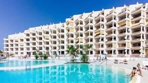 Een hotel met een groot zwembad, meerdere balkons en een helderblauwe lucht.