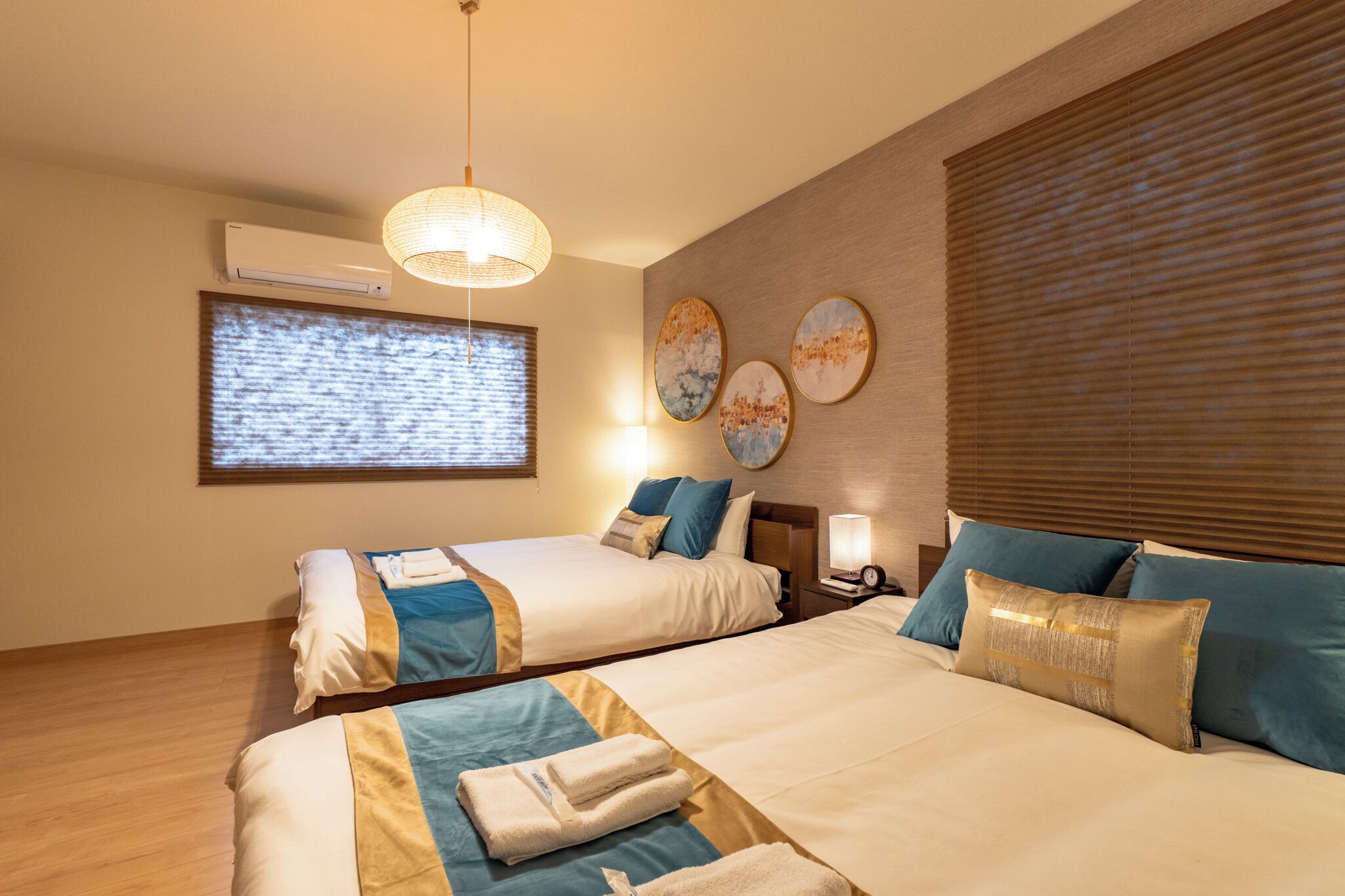 Une chambre d’hôtel avec deux lits, une grande fenêtre avec stores, un climatiseur fixé au mur et un luminaire au plafond.