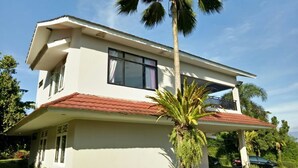 Een tweelaags huis met een rode dakbedekking, een balkon op de bovenverdieping en een verzorgde tuin met palmboompjes.