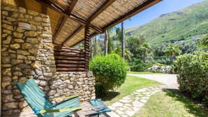 Un bâtiment en pierre doté d’un patio au toit de chaume, de deux fauteuils de jardin verts et d’un chemin en pierre menant à un jardin luxuriant, agrémenté de palmiers et ayant une chaîne de montagnes en toile de fond.