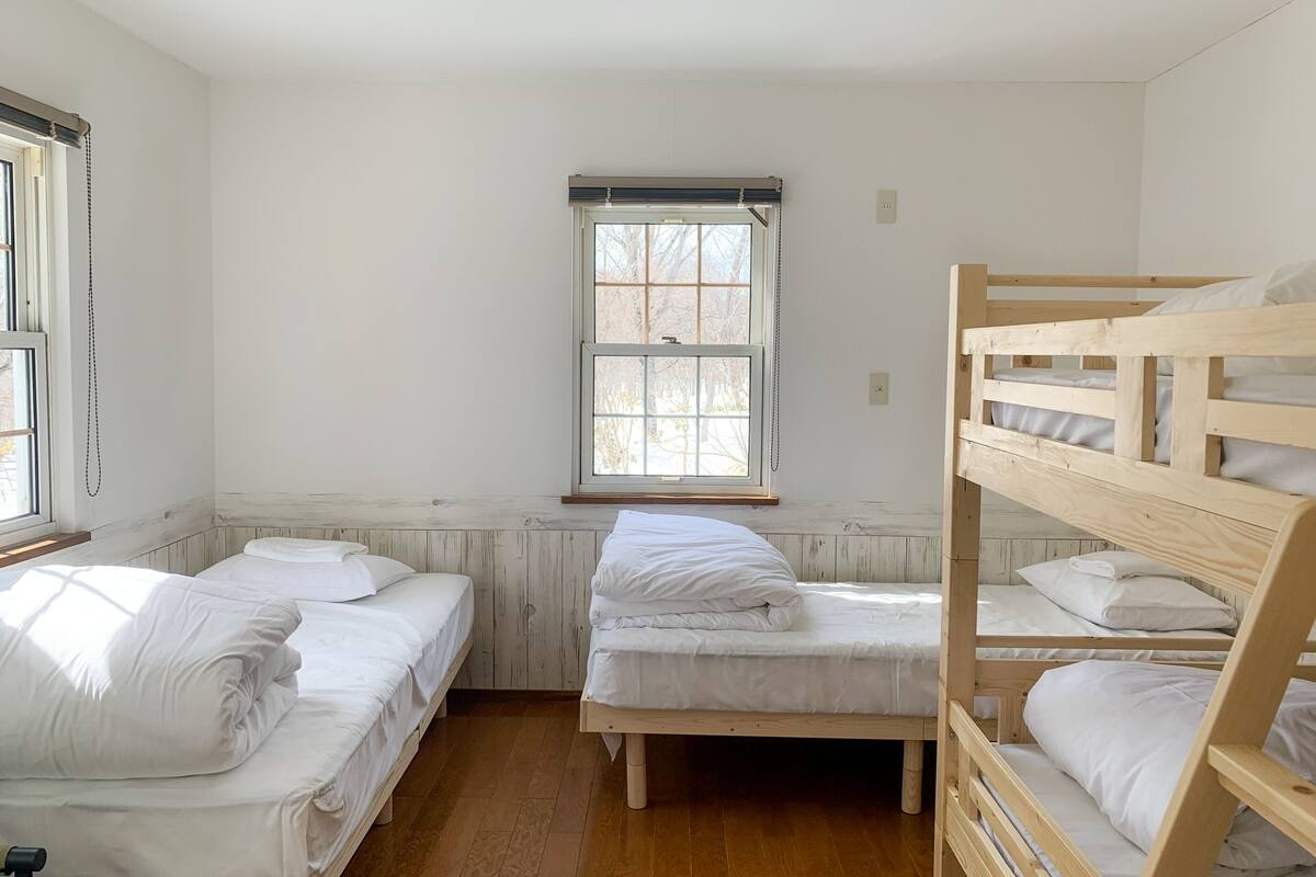 Quarto de estudantes com duas camas individuais e uma cama de dossel, piso de madeira, janela com persianas e radiador.