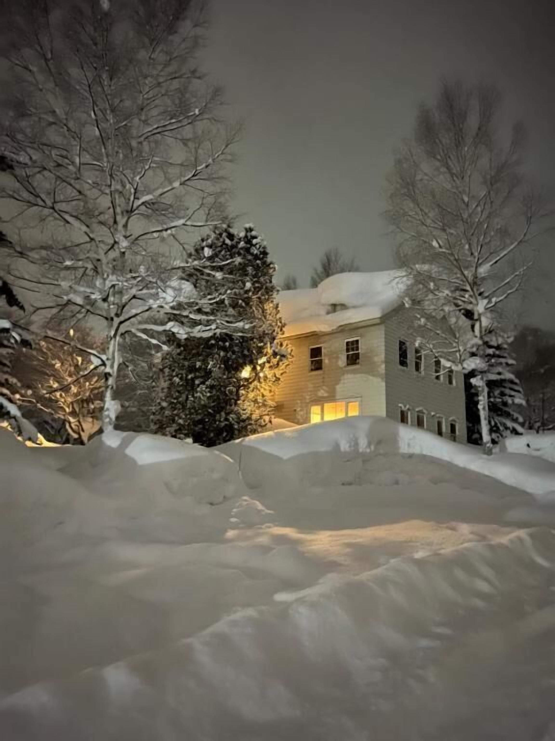 A house surrounded by snow with a visible front porch and a lit window.
