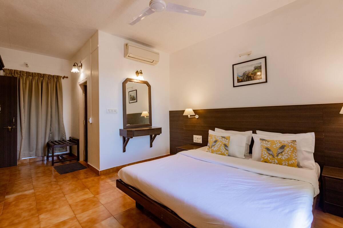 Habitación de hotel con una cama grande, un ventilador de techo, un aire acondicionado de pared, una mesita de noche con lámpara y un cuadro enmarcado en la pared.