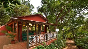 A red-roofed building with a covered patio area, surrounded by lush greenery and a paved walkway. - Radha Cottage (Karjat)