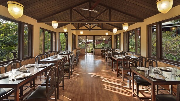 A spacious dining area with wooden tables and chairs, large windows, and hanging lights. - Radha Cottage (Karjat)