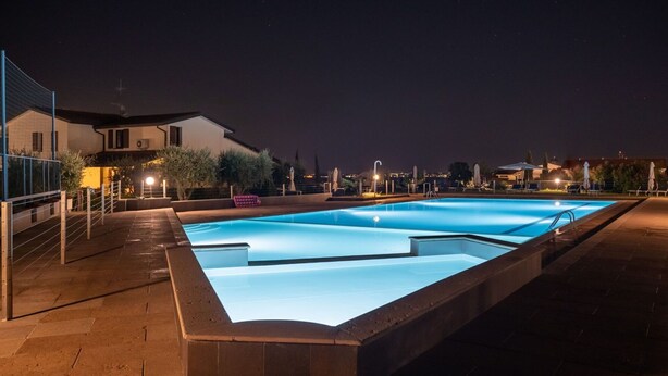 Une piscine rectangulaire, bordée d’une terrasse carrelée, entourée d’une clôture, et un bâtiment avec un balcon en arrière-plan.