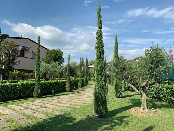 A well-maintained garden with a paved pathway, neatly trimmed hedges, and tall evergreen trees.