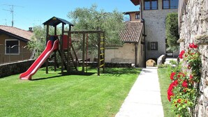 A playground with a red slide and climbing structures, a stone building with a tiled roof, and a pathway leading to a house.