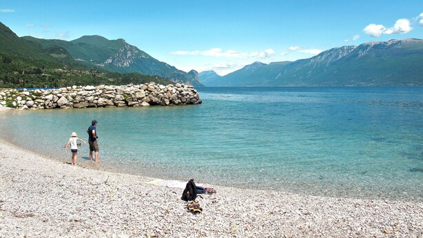 A pebbled beach with clear water, rocky shore, and mountains in the background.