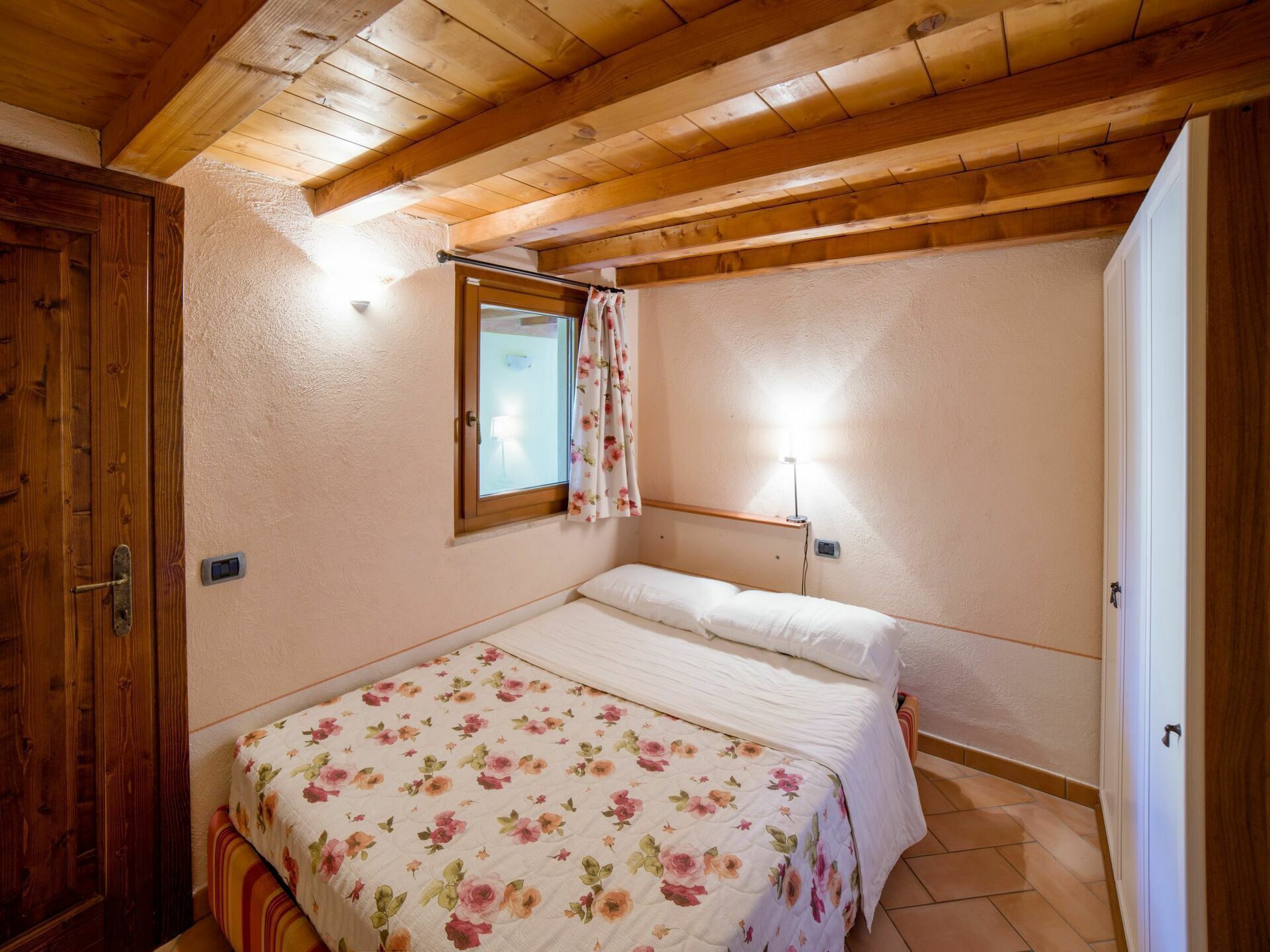 Un dormitorio con techo de madera, cama con ropa de cama floral, ventana con cortinas y una lámpara de noche.