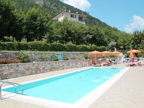 Une piscine entourée de chaises longues et de parasols, elle-même bordée d’un mur de pierre et de verdure, avec une montagne en toile de fond.