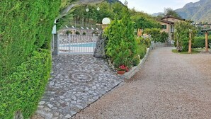 Un vialetto conduce a un'area piscina con ingresso in pietra, immersa nel verde e con le montagne sullo sfondo.