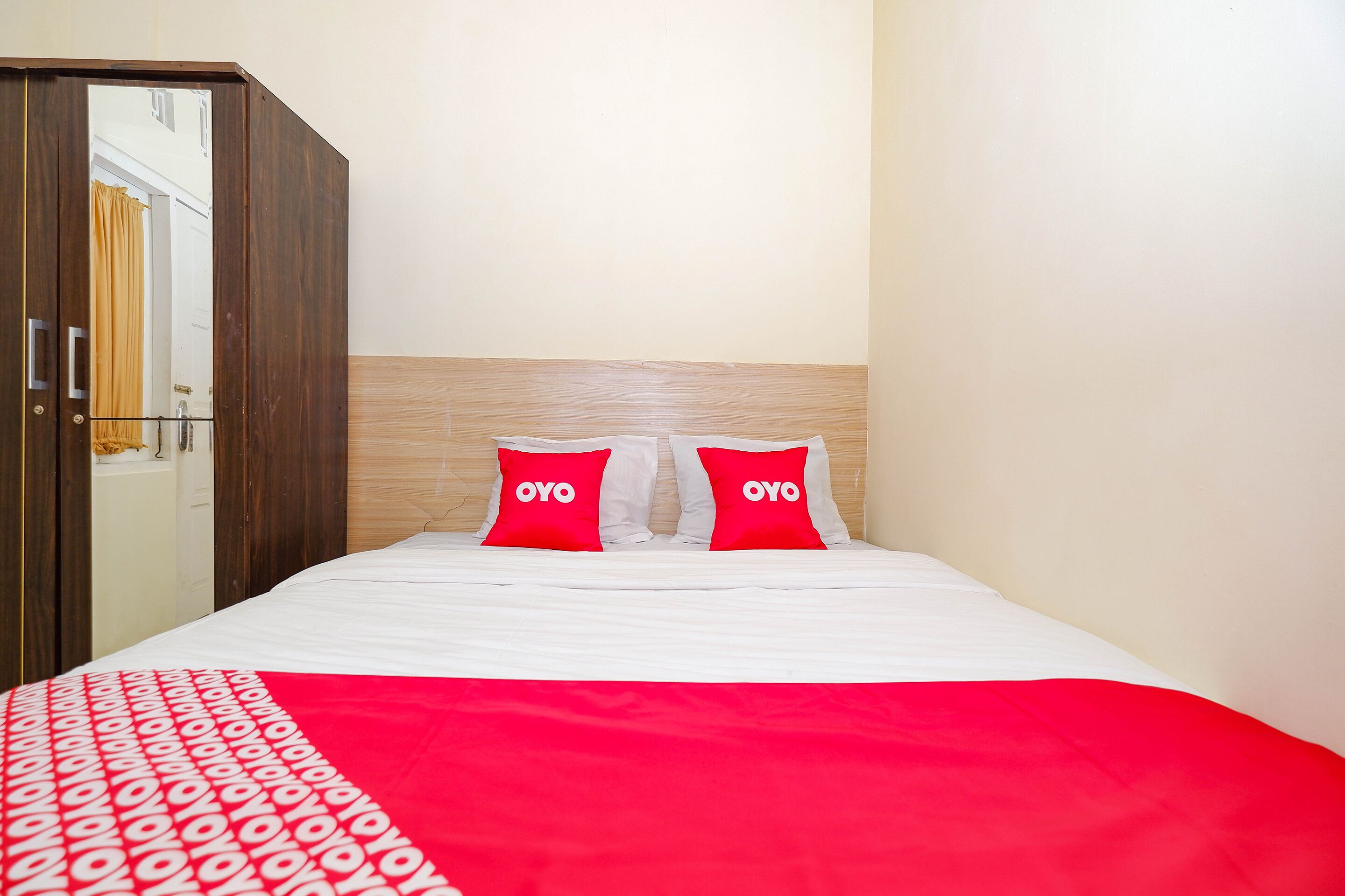 Een hotelkamer met een bed, twee rode kussens met de tekst 'OYO', een houten garderobe en een witte muur.