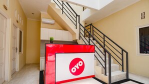 A hotel lobby with a staircase, a reception desk with a red and white design, and a plant on the desk. - OYO 2856 Wisma Fortuna Syariah (Surakarta)