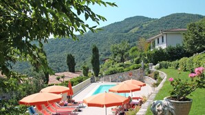 Ein Poolbereich mit mehreren Liegestühlen und Sonnenschirmen, umgeben von viel Grün und Blick auf die Berge.