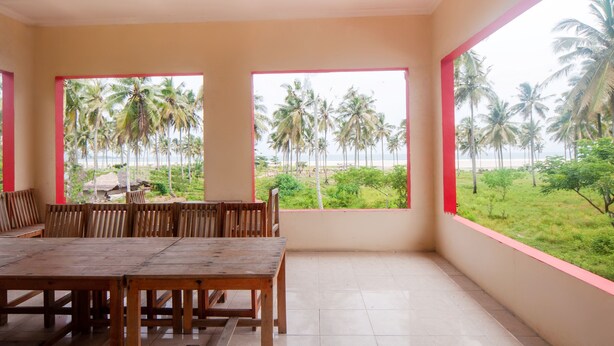 A room with a wooden table and chairs, large windows with a view of palm trees and a beach. - OYO 2471 Swarna Raya (Bayah)