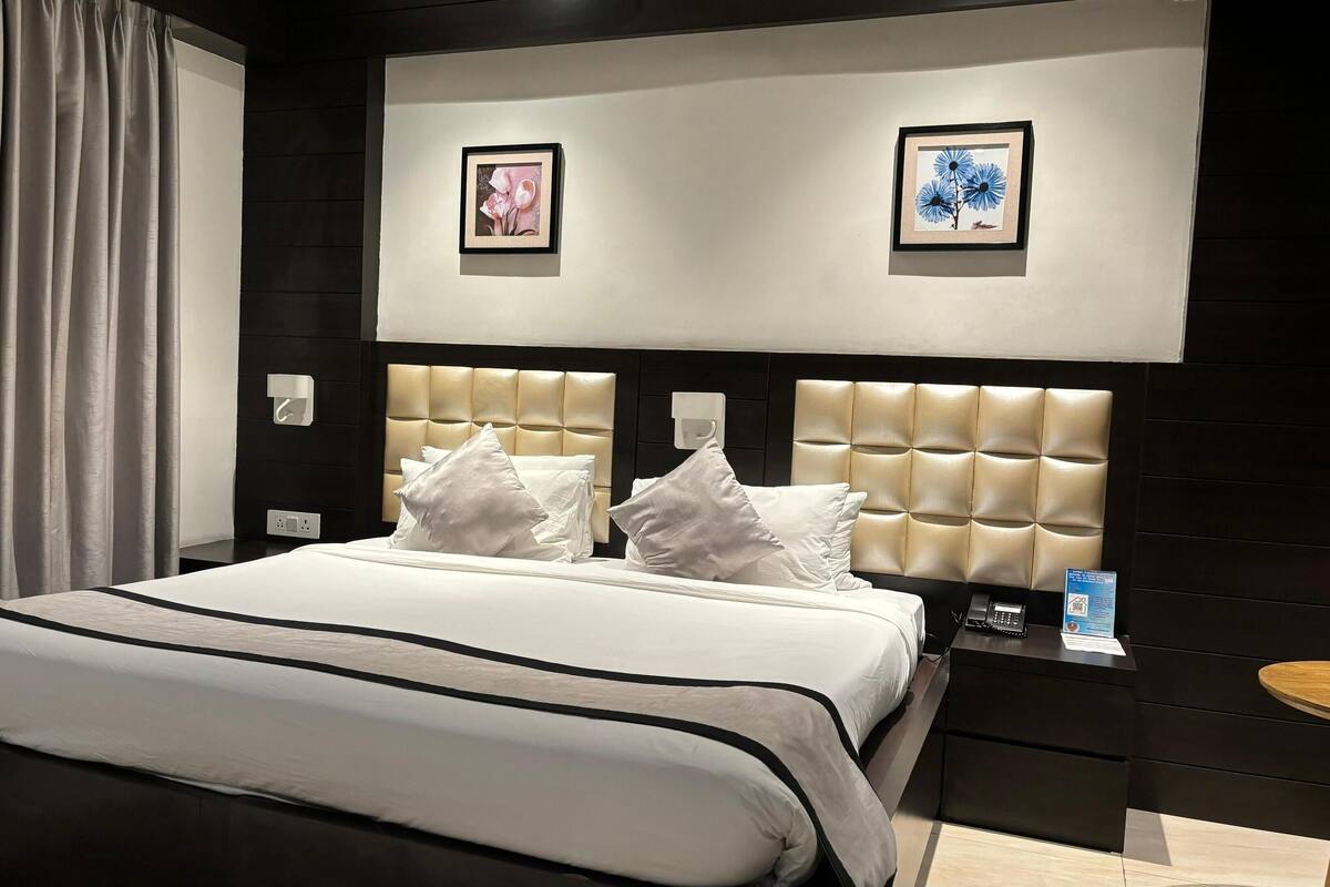 A modern hotel room with a large bed, two framed pictures on the wall, a bedside table with a phone, and a small round table.