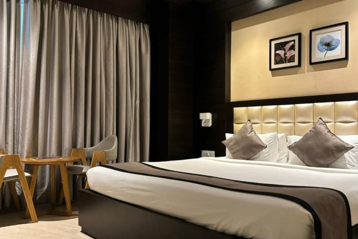 A modern hotel room with a large bed, a small table, two chairs, and a large window with curtains.
