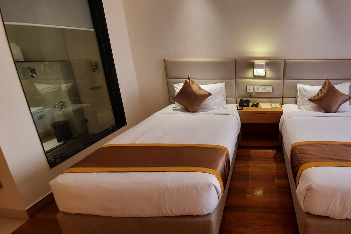 A hotel room with two beds, a bedside table with a lamp, a mirror, and a window with curtains.