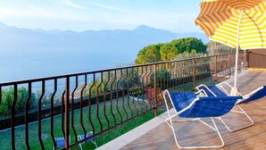 A balcony with a view of the sea and mountains, two blue lounge chairs, and a striped umbrella.