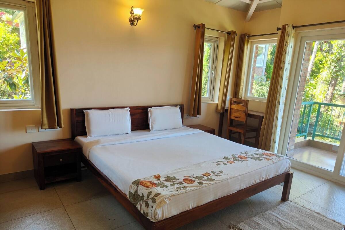 A bedroom with a large bed, a nightstand, a chair, a balcony with a railing, and a wall-mounted light fixture.