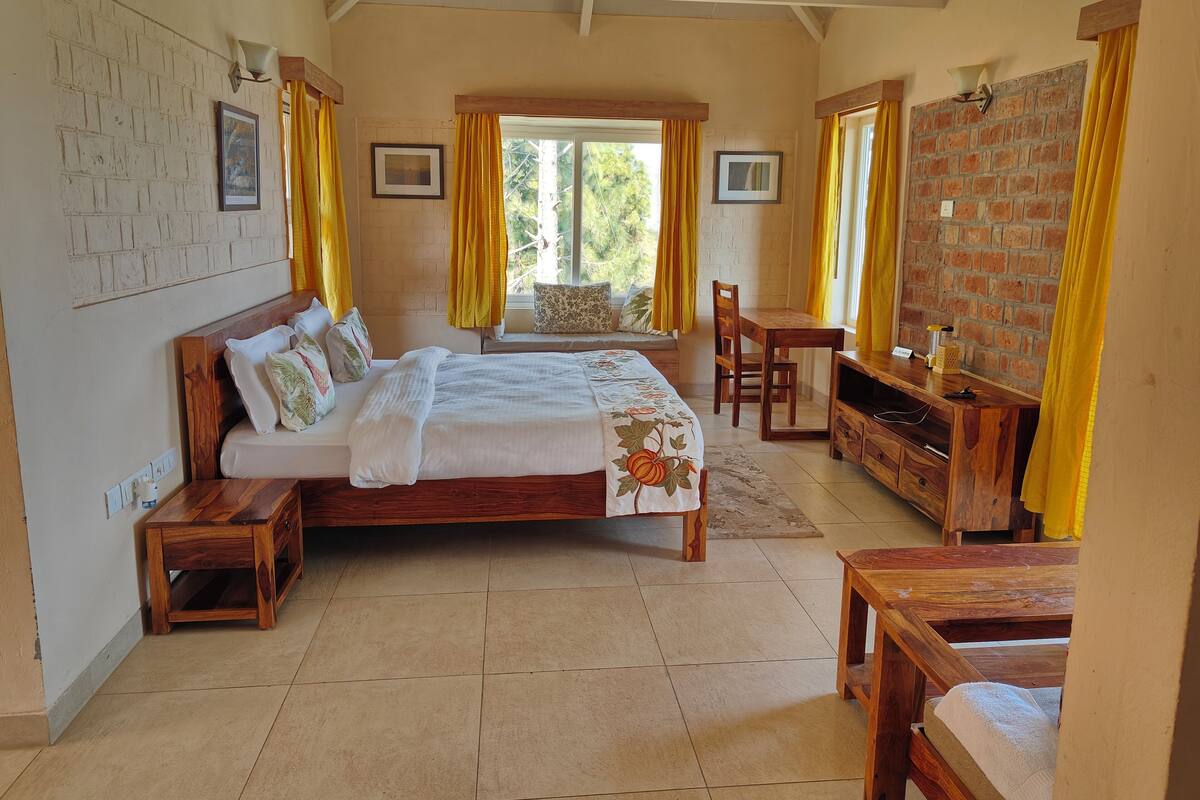 A bedroom with a large bed, a wooden desk, a chair, and a TV. There are windows with curtains, a brick wall, and a ceiling with wooden beams.