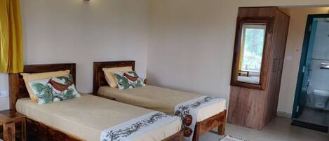 A room with two wooden beds, a small wooden table, a window with a view, and a bathroom door.