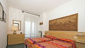 Hotellihuone, jossa on kaksi sänkyä, lipasto, peili, ikkuna, jossa on läpikuultavat verhot, ja suuri seinävaate seinällä.