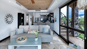 A modern living room with a sofa, coffee table, and a view of the kitchen and outdoor area.