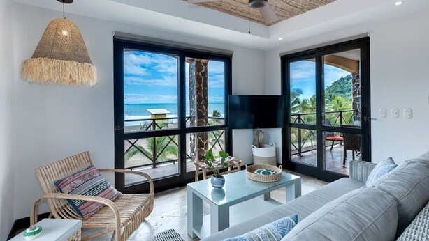 A spacious living room with a large window offering a view of the ocean, a ceiling fan, and a woven pendant light.