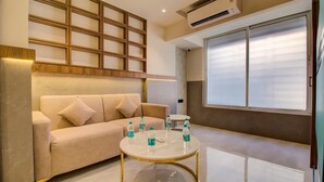 A modern living room with a beige sofa, a glass coffee table with water bottles, and a large window with blinds. - Hotel Malad Inn (Mumbai)