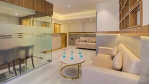 A modern hotel room with a glass partition, a round gold table with water bottles, a beige sofa, and a wooden bookshelf. - Hotel Malad Inn (Mumbai)