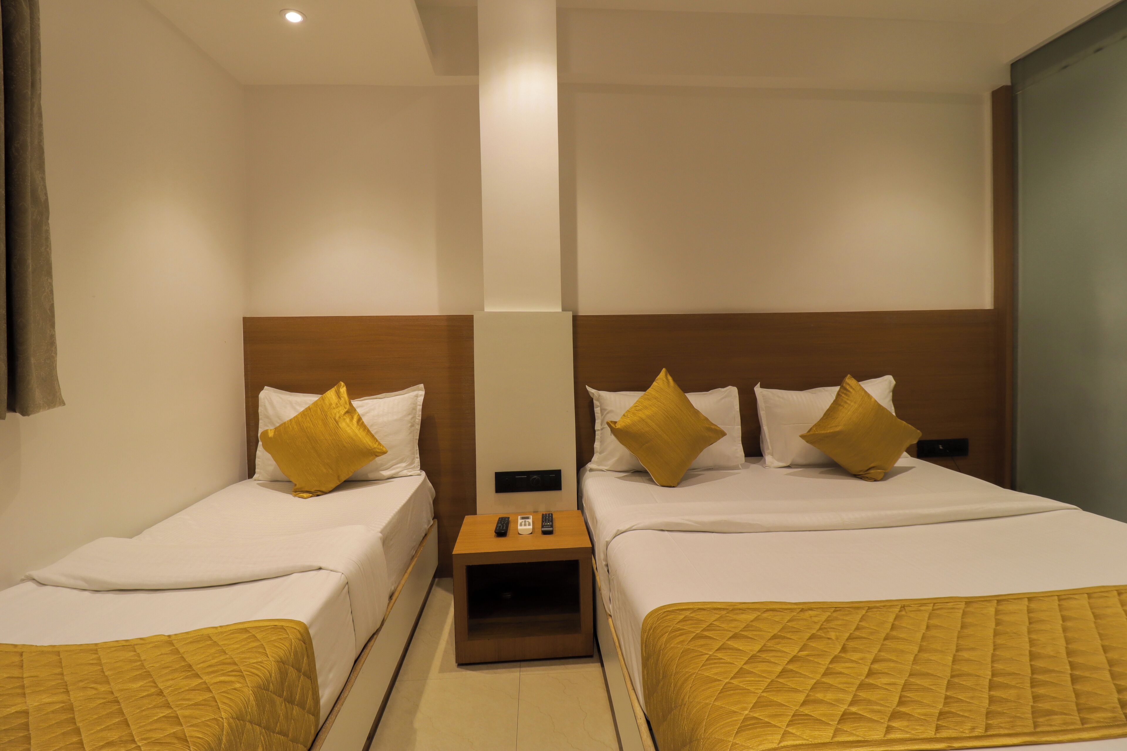 A hotel room with two single beds, each with a wooden headboard, white bedding, and a yellow quilted throw. There is a small wooden nightstand between the beds with a remote control and a phone on it.