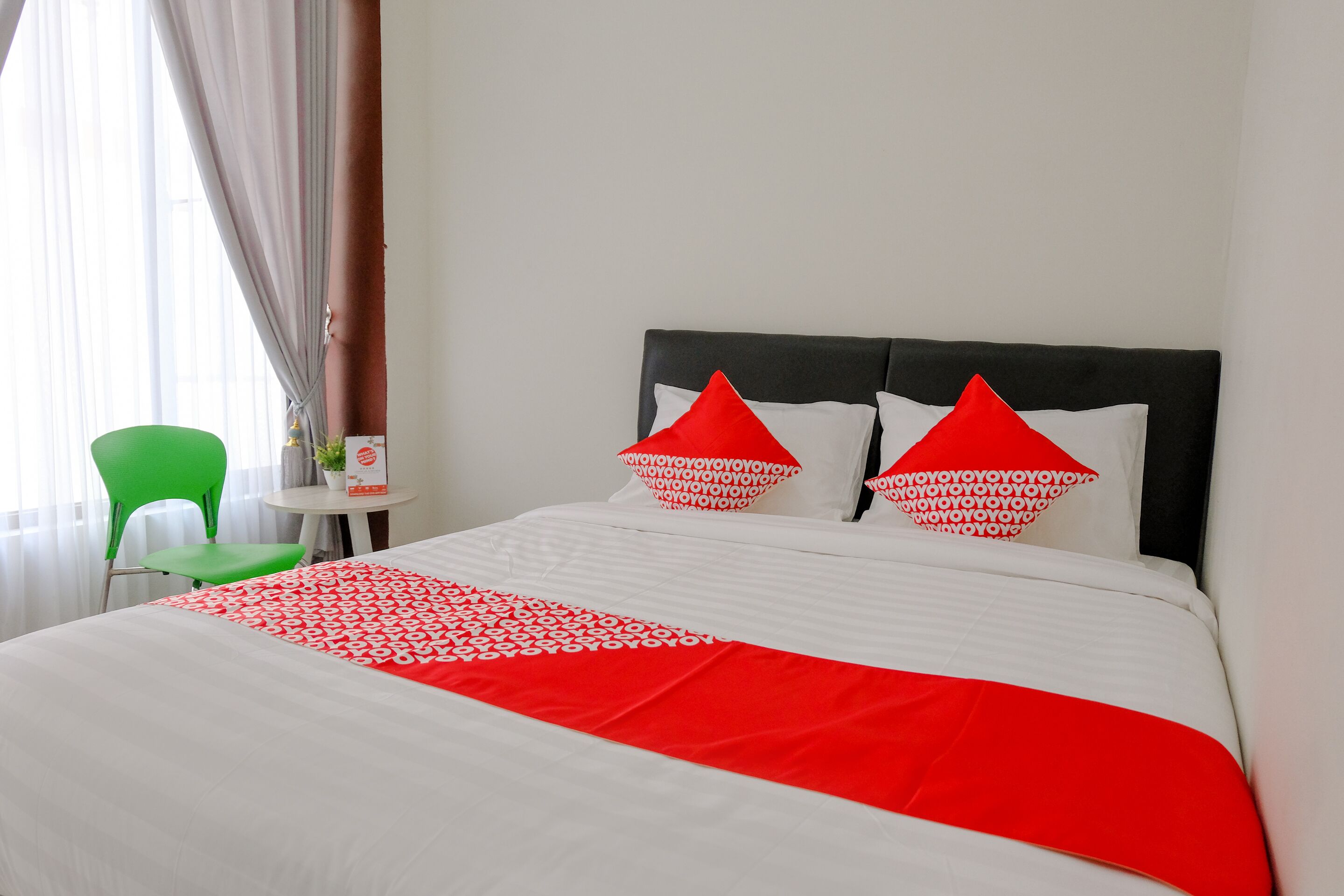 Un lit bien fait, recouvert d’une couvre-lit blanche et rouge, avec deux oreillers rouges et une chaise verte, dans une pièce aux murs blancs et dotée d’une fenêtre avec des rideaux.