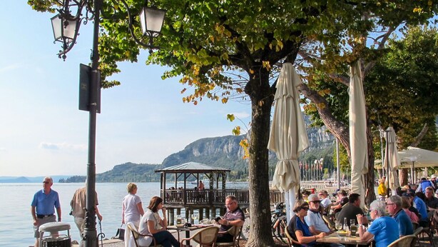 An outdoor dining area with tables and chairs, a covered seating area, and a view of a lake with mountains in the background. - Cosy apartment for 4 guests with WIFI, pool, TV, patio and pets allowed (Beati Bassi)