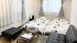 Una habitación de hotel con dos camas, una mesita con lámpara, un taburete negro y una ventana con cortinas estampadas.