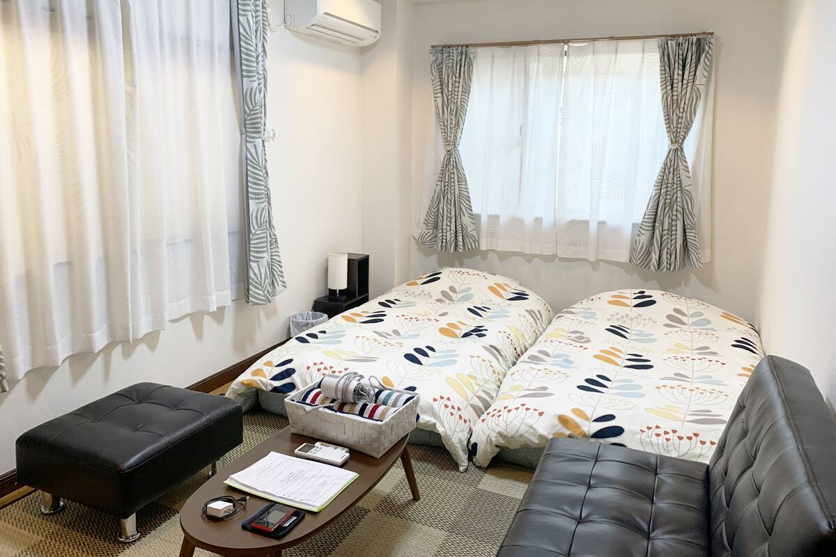 Una habitación de hotel con dos camas, una mesita con lámpara, un taburete negro y una ventana con cortinas estampadas.
