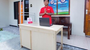 A man in a red shirt standing in front of a desk with a laptop, a wooden chair, and a sign on the wall. - Hotel O Griya Annisa (Tegal)
